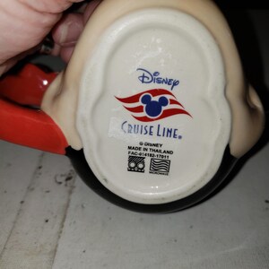 May include: A Disney Cruise Line mug with a black base, red handle, and a cream-colored top. The mug features the Disney Cruise Line logo and text on the bottom. The mug is microwave safe.