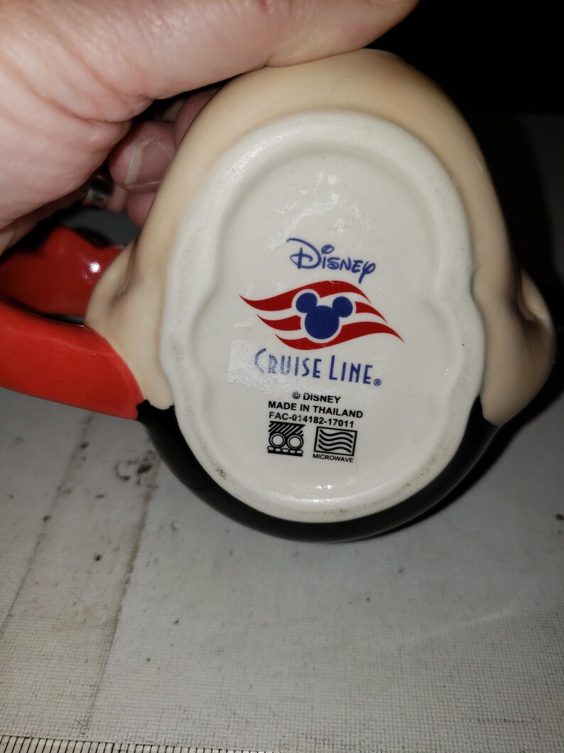 May include: A Disney Cruise Line mug with a black base, red handle, and a cream-colored top. The mug features the Disney Cruise Line logo and text on the bottom. The mug is microwave safe.