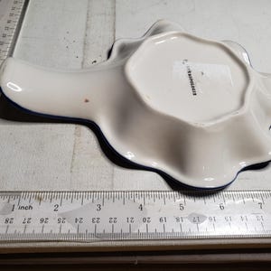May include: White ceramic spoon rest with a wavy edge and a dark blue trim. The spoon rest is approximately 6 inches long. The underside has the text "ANTHROPOLOGIE".