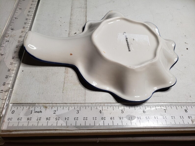May include: White ceramic spoon rest with a wavy edge and a dark blue trim. The spoon rest is approximately 6 inches long. The underside has the text "ANTHROPOLOGIE".
