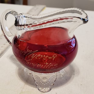 May include: A small, clear glass pitcher with a red interior and three legs. The pitcher has a handle and is engraved with the text "Mary Meyers 9 Mayville".