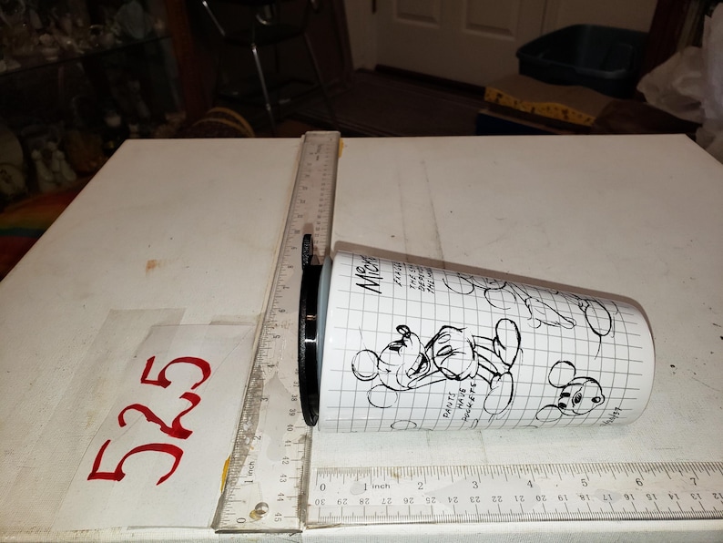 May include: White tumbler with black Mickey Mouse illustrations and text. The tumbler has a black lid and is measured with a ruler. The text on the tumbler includes "Pants have pockets".