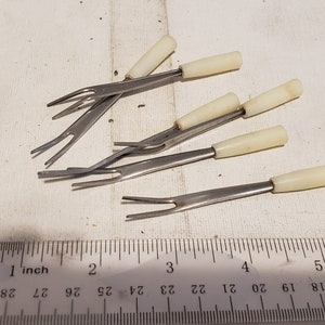 May include: A collection of six small, stainless steel forks with off-white handles. The forks have two tines and are arranged on a white surface next to a ruler. Each fork measures approximately 5 inches in length.