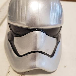 May include: A silver-colored First Order Stormtrooper helmet with black visor and face details. The helmet has vertical ridges on the top and a smooth, metallic finish. A Star Wars collectible.