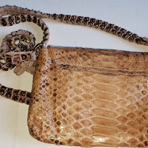 May include: A small, light brown snakeskin purse with a beaded strap. The purse has a flap closure and a small, rectangular shape.