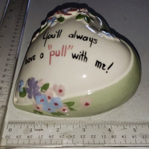May include: A white heart-shaped ceramic trinket box with a green rim and floral designs. The box has a message that reads "You'll always have a "pull" with me!"