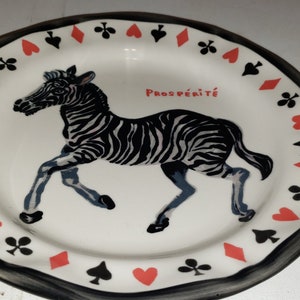 May include: A white ceramic plate with a black and white zebra illustration and the word "Prosp&eacute;rit&eacute;" in red. The plate has a black border with a repeating pattern of red hearts, black spades, and red diamonds.