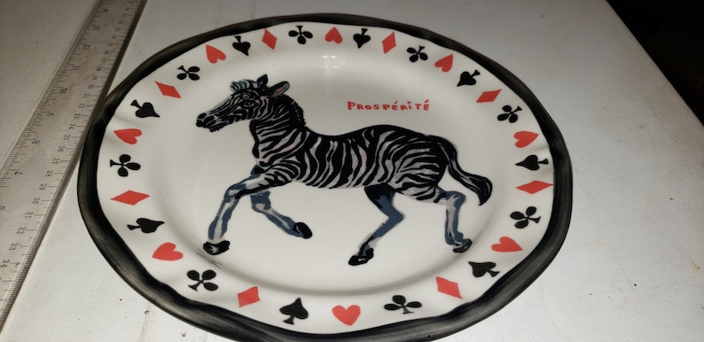 May include: A white ceramic plate with a black and white zebra illustration and the word "Prosp&eacute;rit&eacute;" in red. The plate has a black border with a repeating pattern of red hearts, black spades, and red diamonds.