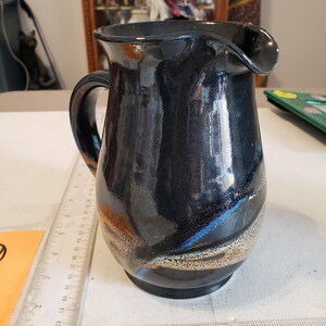 May include: A black ceramic pitcher with a curved spout and handle. The pitcher has a glossy finish with streaks of blue, brown, and gold. The pitcher is approximately 6 inches tall.