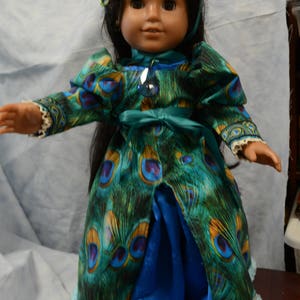 May include: A doll wearing a blue and green peacock-patterned dress with a teal sash and a blue satin skirt. The doll is wearing a crown with blue and green jewels.
