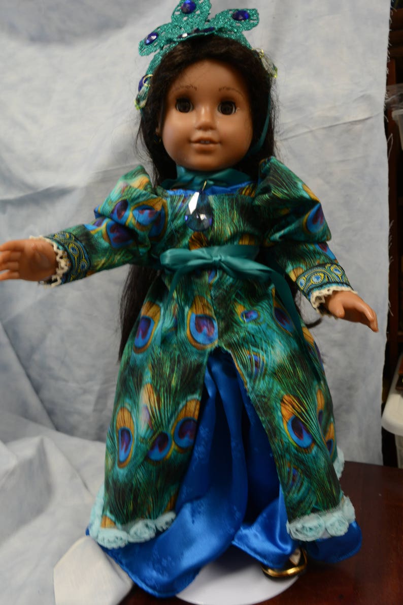 May include: A doll wearing a blue and green peacock-patterned dress with a teal sash and a blue satin skirt. The doll is wearing a crown with blue and green jewels.