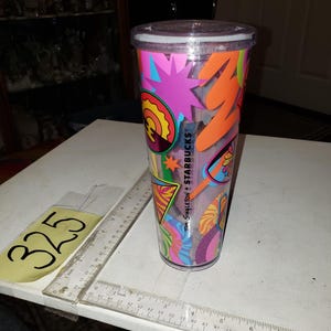 May include: A clear plastic reusable cup with a colorful design featuring geometric shapes, stars, and the text "Tim Singleton + Starbucks".