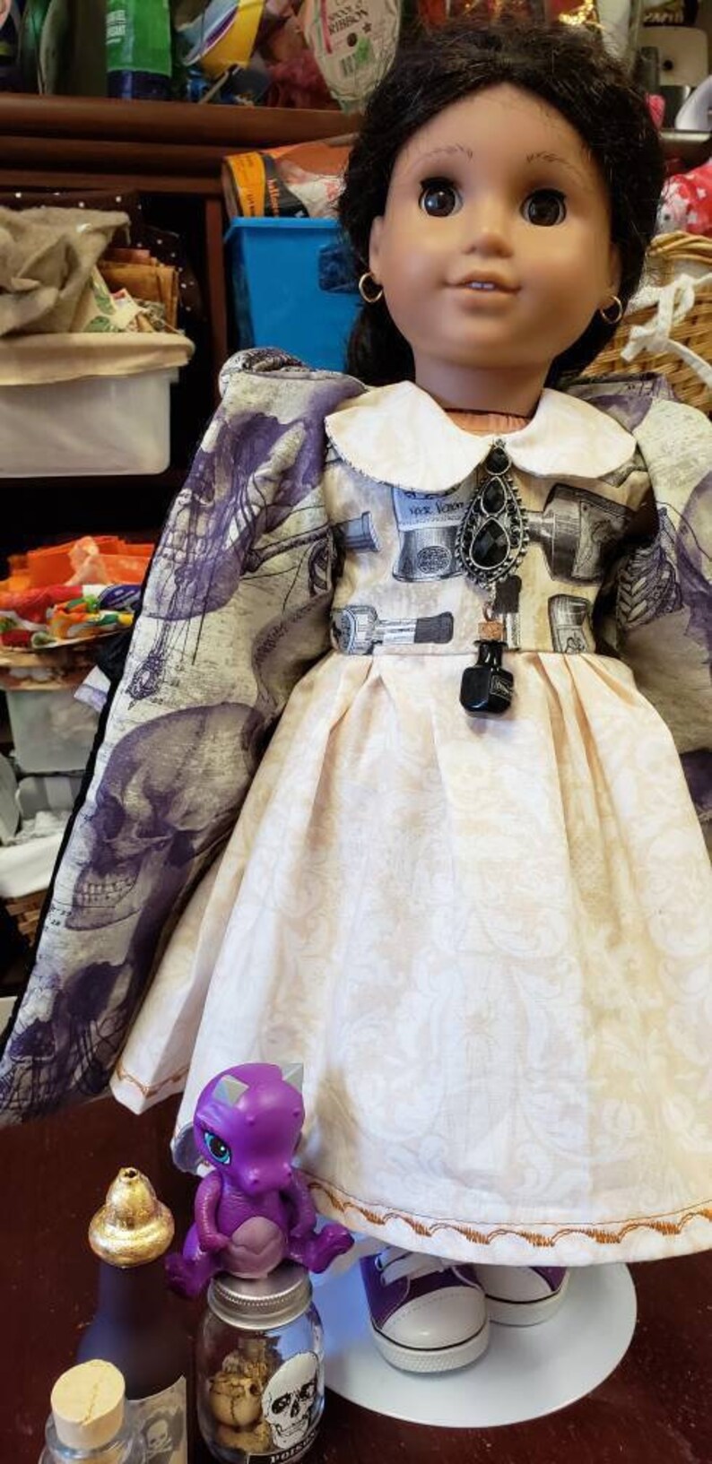 May include: A doll wearing a cream-colored dress with a white collar and a patterned bodice. The doll has dark hair and is accessorized with a black pendant. A purple toy dragon sits on a jar with a skull label.