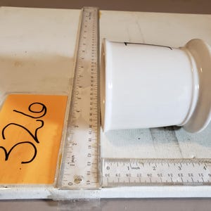 以下が含まれることがあります： 丸みを帯びたベースとわずかにフレアした上部を備えた白いセラミック容器。容器の横には定規が置かれ、センチメートル単位の測定値が表示されています。数字「326」の黄色い付箋も表示されています。