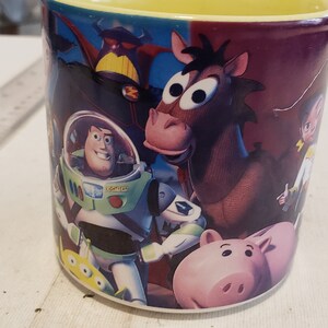 May include: A ceramic coffee mug with a colorful image of characters from the Toy Story movie franchise. The characters include Woody, Buzz Lightyear, and Hamm. The mug has a yellow rim.