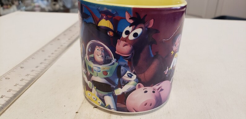 May include: A ceramic coffee mug with a colorful image of characters from the Toy Story movie franchise. The characters include Woody, Buzz Lightyear, and Hamm. The mug has a yellow rim.