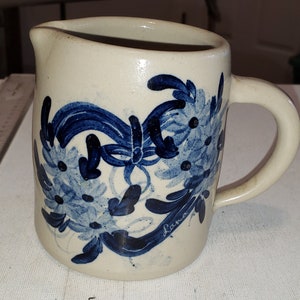 May include: A white ceramic pitcher with a blue floral design. The pitcher has a handle and a spout. The floral design features blue flowers and leaves.