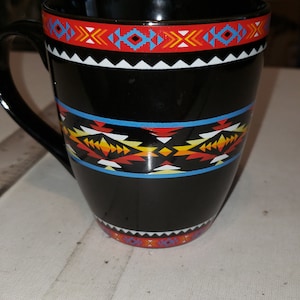 May include: Black ceramic mug with a colorful geometric pattern in red, orange, yellow, blue, and white. The pattern is reminiscent of Native American art.
