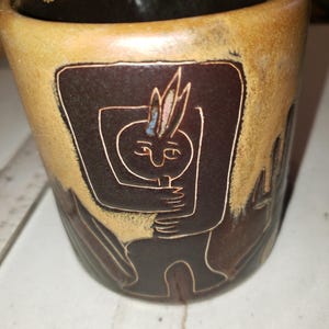 May include: A ceramic mug with a brown and tan color scheme. The mug features a dark brown rectangular design with a white outline of a person holding an object. The mug has a dark brown interior.