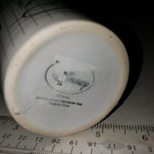 May include: White ceramic Disney mug with black sketched design. The bottom of the mug has a Disney logo and text. A ruler is placed next to the mug for scale.