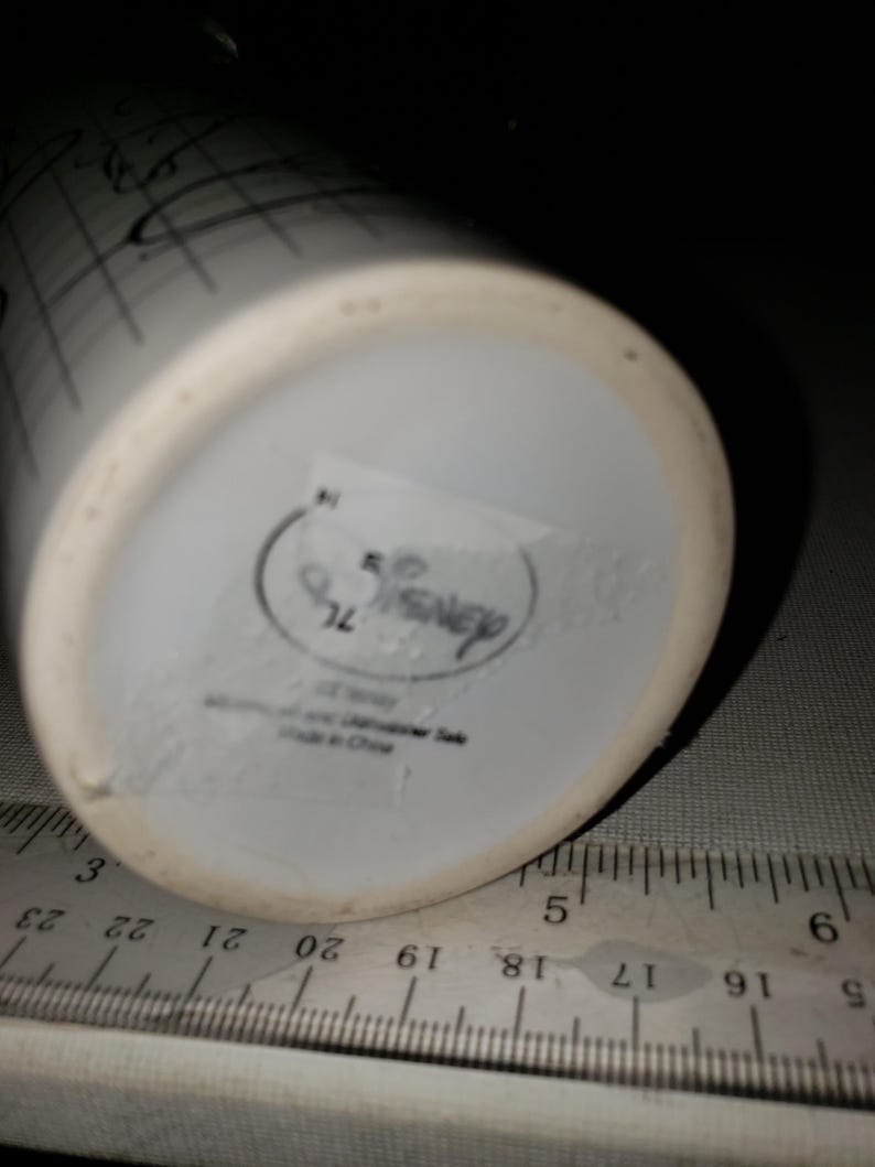 May include: White ceramic Disney mug with black sketched design. The bottom of the mug has a Disney logo and text. A ruler is placed next to the mug for scale.