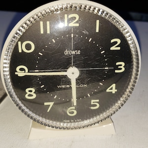 May include: White alarm clock with a black face and silver trim. The clock face has black numbers and hands, and the word "drowse" printed in white. The clock is made by Westclox and says "Made in U.S.A."