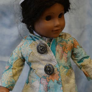May include: A doll wearing a white and blue jacket with a world map print and compass buttons. The doll has brown hair and is wearing white sunglasses.