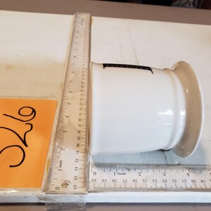 May include: A white ceramic container with a wide rim and a slightly tapered body. The container has a black marking on the side. A ruler is placed next to the container, showing measurements in inches.