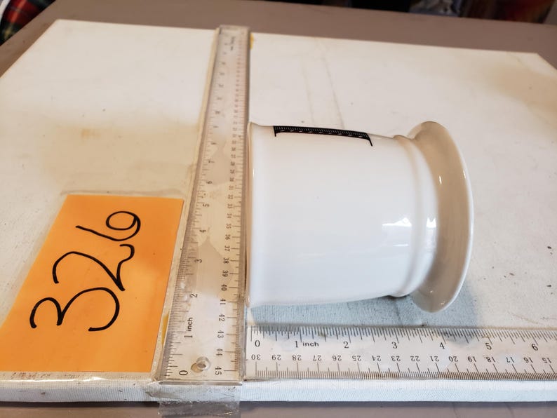May include: A white ceramic container with a wide rim and a slightly tapered body. The container has a black marking on the side. A ruler is placed next to the container, showing measurements in inches.