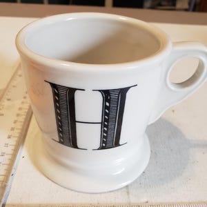May include: White ceramic mug with a black letter "H" in a decorative font. The mug has a rounded base and a handle. A ruler is placed next to the mug, showing inches.
