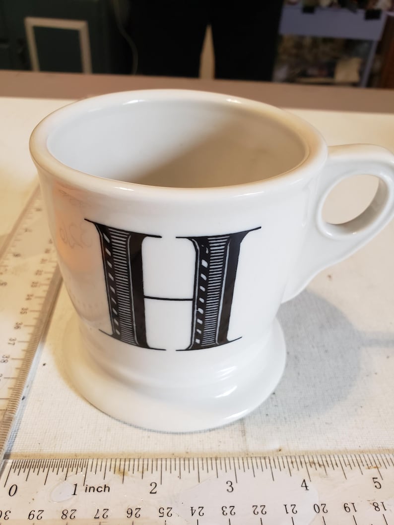 May include: White ceramic mug with a black letter "H" in a decorative font. The mug has a rounded base and a handle. A ruler is placed next to the mug, showing inches.