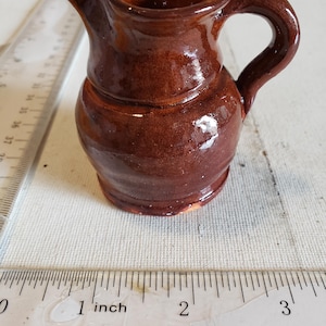May include: A small, handmade brown ceramic pitcher with a handle and spout. The pitcher is approximately 2 inches tall. The glaze has a glossy finish. The pitcher is sitting on a white surface next to a ruler.