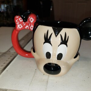 May include: A ceramic mug shaped like Minnie Mouse's face. The mug features a red handle and bow with white anchor designs. The mug has black ears, eyelashes, and a black mouth. The background is a light-colored surface.