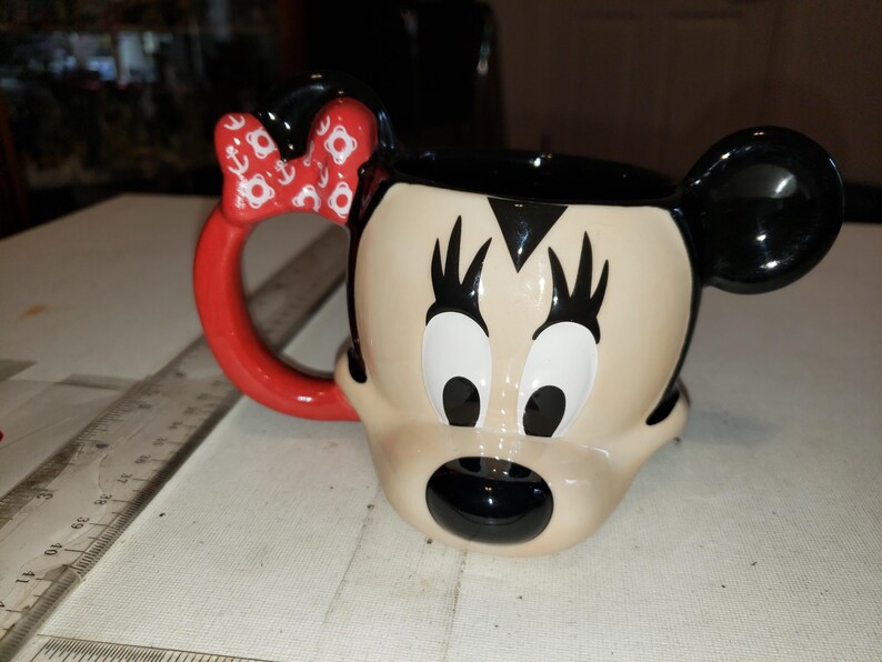 May include: A ceramic mug shaped like Minnie Mouse's face. The mug features a red handle and bow with white anchor designs. The mug has black ears, eyelashes, and a black mouth. The background is a light-colored surface.