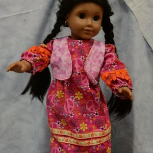 May include: A doll with long black braids wearing a pink dress with a floral and peace sign pattern. The dress has a pink vest with a white and pink tie-dye pattern. The doll is wearing pink shoes.