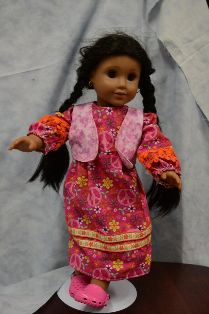 May include: A doll with long black braids wearing a pink dress with a floral and peace sign pattern. The dress has a pink vest with a white and pink tie-dye pattern. The doll is wearing pink shoes.