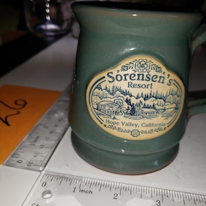 May include: A sage green ceramic mug with a handle, featuring a cream-colored oval decal with a blue illustration of buildings and trees. The text reads "Sorensen's Resort" and "Hope Valley, California". A ruler shows the mug is 3 inches wide.