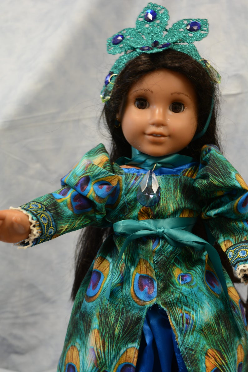 May include: A doll wearing a teal and green peacock-patterned dress with a teal sash and a crown with blue jewels. The doll has long black hair and brown eyes.