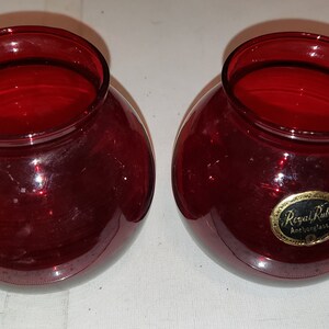 May include: Two round, ruby red glass bowls with wide openings. One bowl has a gold and black label that reads "Royal Ruby". The bowls are likely for decorative use or as small planters.