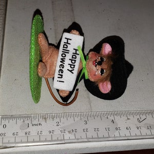 K&ouml;nnte beinhalten: Eine Halloween-Dekoration mit einer Maus, die ein Schild mit der Aufschrift "Happy Halloween!" h&auml;lt. Die Maus hat einen schwarzen Hut, rosa Ohren und einen braunen K&ouml;rper. Sie h&auml;lt einen gr&uuml;nen, glitzernden Gegenstand. Die Dekoration ist etwa 7.6 cm lang.