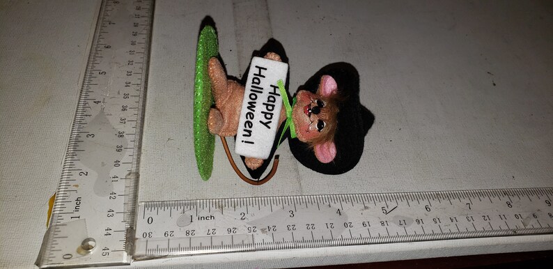 K&ouml;nnte beinhalten: Eine Halloween-Dekoration mit einer Maus, die ein Schild mit der Aufschrift "Happy Halloween!" h&auml;lt. Die Maus hat einen schwarzen Hut, rosa Ohren und einen braunen K&ouml;rper. Sie h&auml;lt einen gr&uuml;nen, glitzernden Gegenstand. Die Dekoration ist etwa 7.6 cm lang.