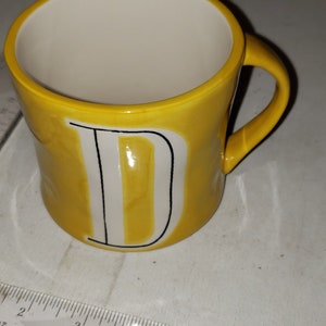 Op de afbeelding: Een gele keramische mok met een witte letter "D" aan de voorkant. De mok heeft een zwarte omtrek rond de letter.