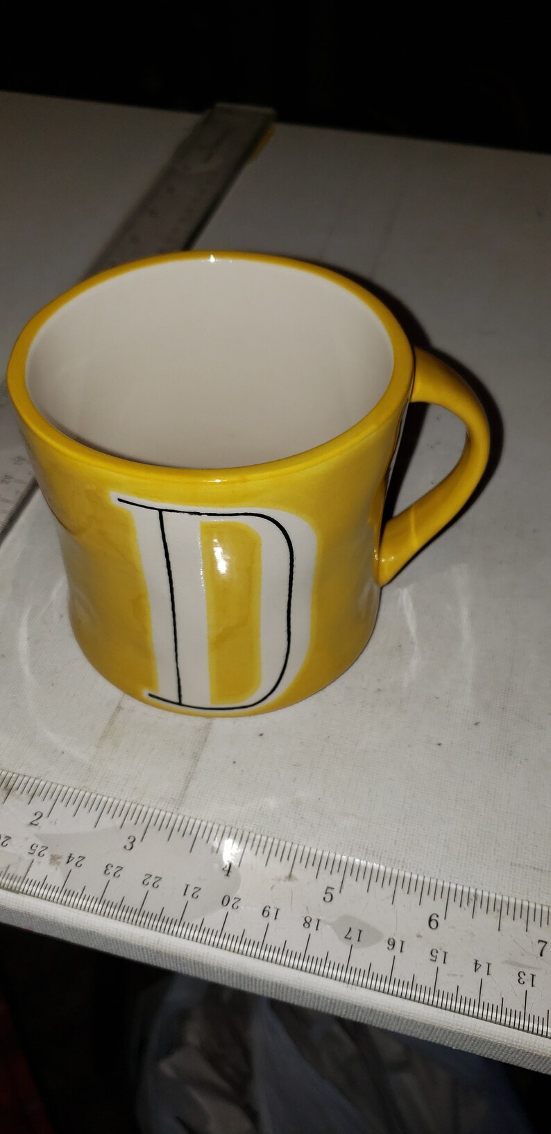 Op de afbeelding: Een gele keramische mok met een witte letter "D" aan de voorkant. De mok heeft een zwarte omtrek rond de letter.
