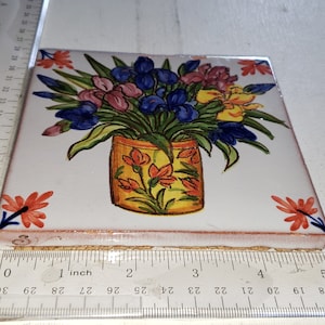 May include: A white ceramic tile with a hand-painted design of a bouquet of flowers in a yellow vase. The flowers are blue, pink, yellow, and orange. The tile is approximately 4 inches by 4 inches.