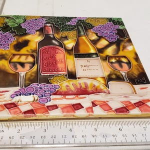 May include: A ceramic tile with a colorful painted design of two wine bottles, two wine glasses, grapes, and a checkered tablecloth. The tile has the word "Ping" written in the bottom right corner.