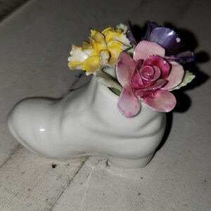 May include: A white ceramic shoe with a bouquet of flowers. The flowers are yellow, pink, and purple.