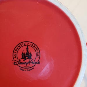 Puede incluir: Una taza de cer&aacute;mica roja con un borde blanco. La base de la taza presenta un logotipo negro de Disney Parks con el texto "AUTHENTIC ORIGINAL" y "Made in Thailand".