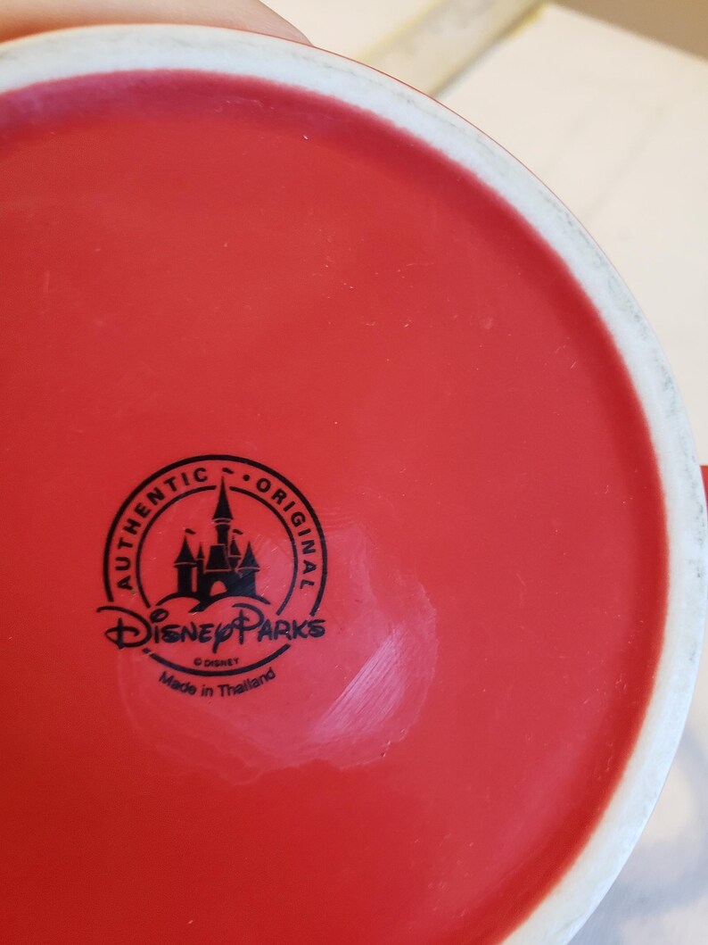 Puede incluir: Una taza de cer&aacute;mica roja con un borde blanco. La base de la taza presenta un logotipo negro de Disney Parks con el texto "AUTHENTIC ORIGINAL" y "Made in Thailand".