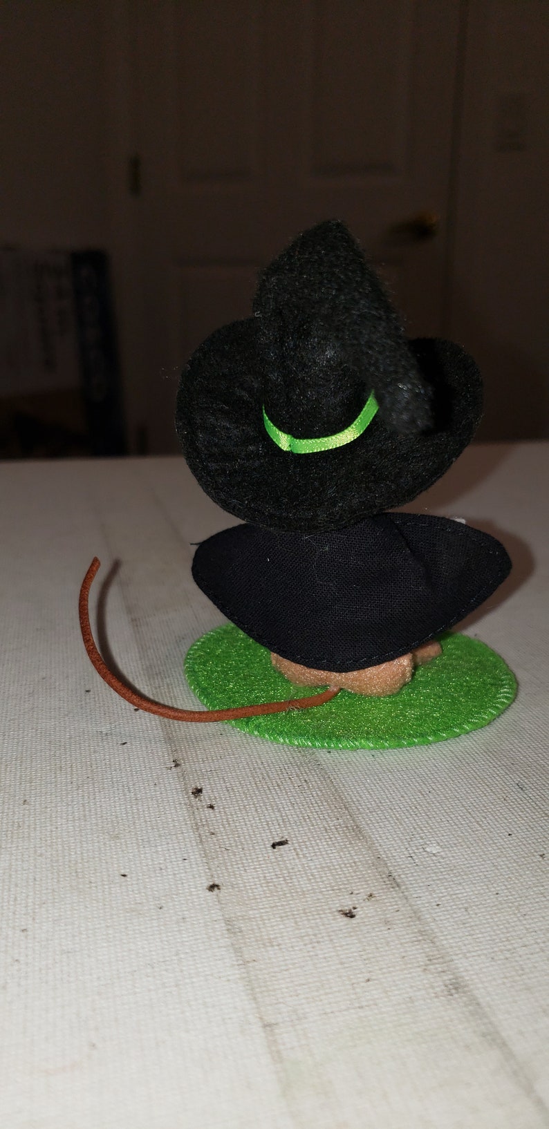 K&ouml;nnte beinhalten: Handgemachte Halloween-Dekoration mit einer kleinen Hexenfigur. Die Hexe hat einen schwarzen Hut mit gr&uuml;nem Band, ein schwarzes Kleid und einen braunen Schwanz. Sie sitzt auf einem gr&uuml;nen Filzkreis.