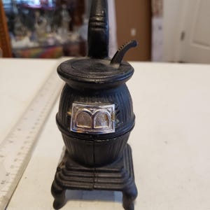 May include: A black cast iron miniature stove with a silver door and a black pipe. The stove is about 4 inches tall and sits on a white surface.
